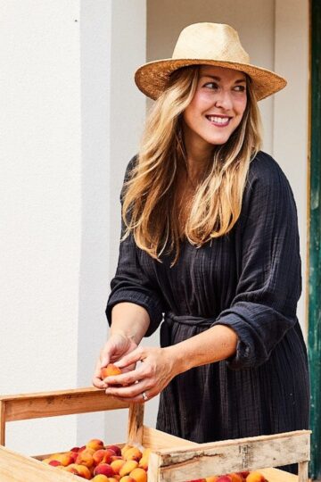 Sarah Copeland stood by a box of fruit, wearing a dark coloured tunic, and straw hat