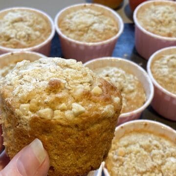 A hand holding an Apple, Oat and Cinnamon Muffin, with more muffins in the background.