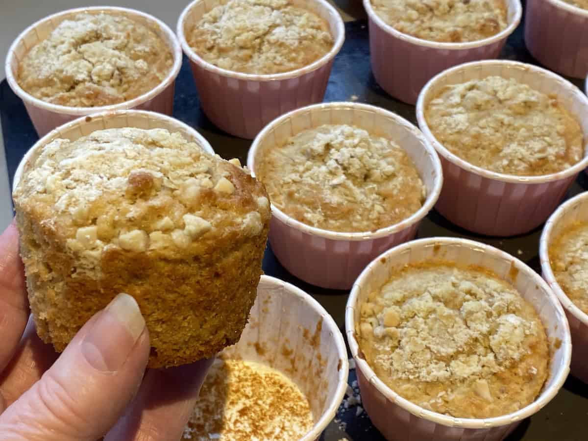 A hand holding an Apple, Oat and Cinnamon Muffin, with more muffins in the background.
