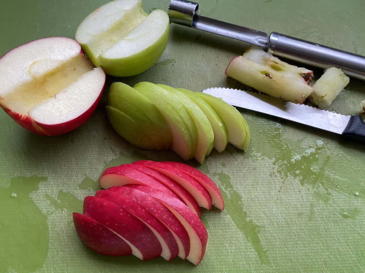 Slices of apple in a bowl of water. Slices of red and green apples on a green chopping board.